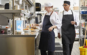 Female sous chef in a white chef beanie seasoning a dish as the head chef in a black chef beanie walks by and checks her work