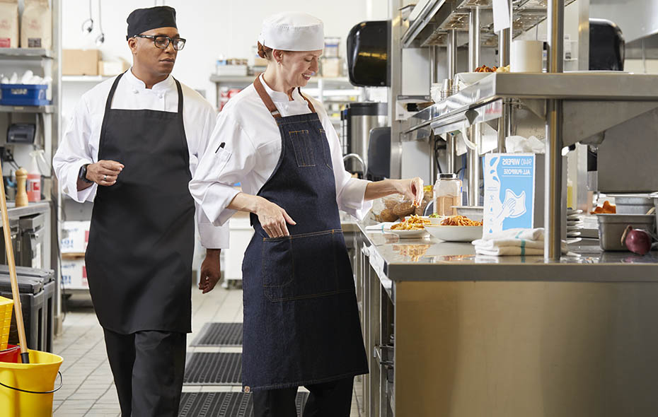 Female sous chef in a white chef beanie seasoning a dish as the head chef in a black chef beanie walks by and checks her work